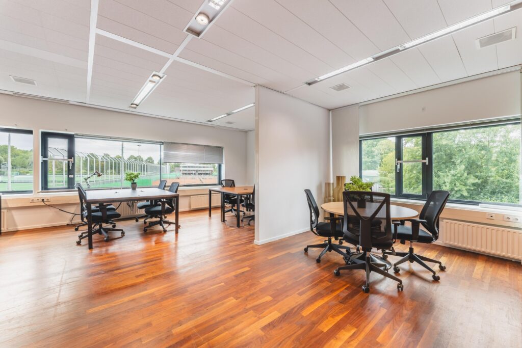 Lichte en moderne kantoorruimte aan de Prof. W.H. Keesomlaan met meerdere bureaus, stoelen en grote ramen met uitzicht op sportvelden en groen.